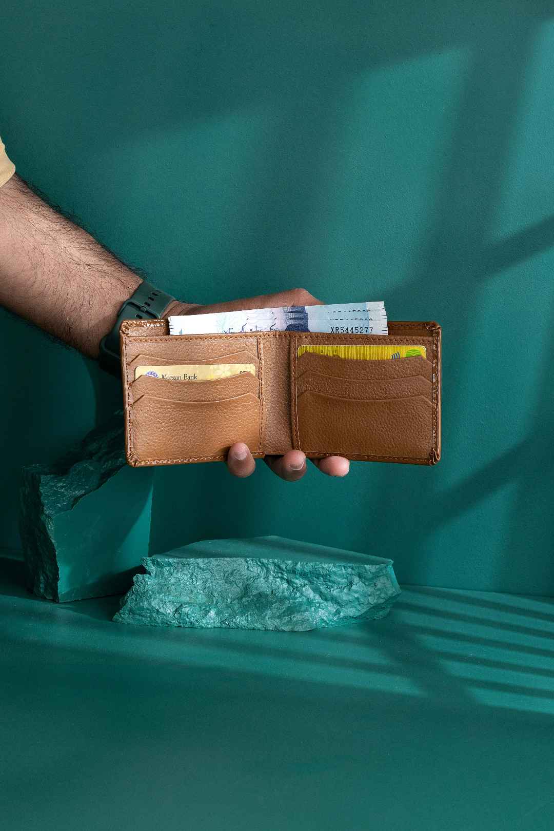 creative studio shot of hand modeling a brown leather wallet with money on a teal background