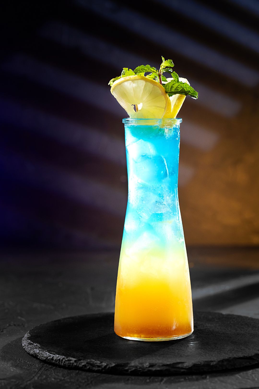Blue and orange layered cocktail with a lemon wedge and mint leaf on a dark surface.
